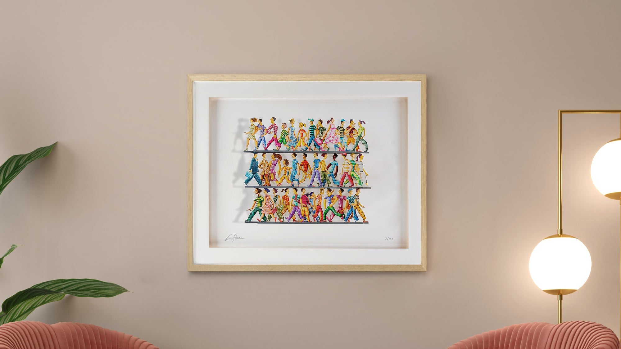 Framed hand-painted papercut artwork by David Gerstein, featuring colorful walking figures, mounted on a neutral wall above two pale pink chairs.