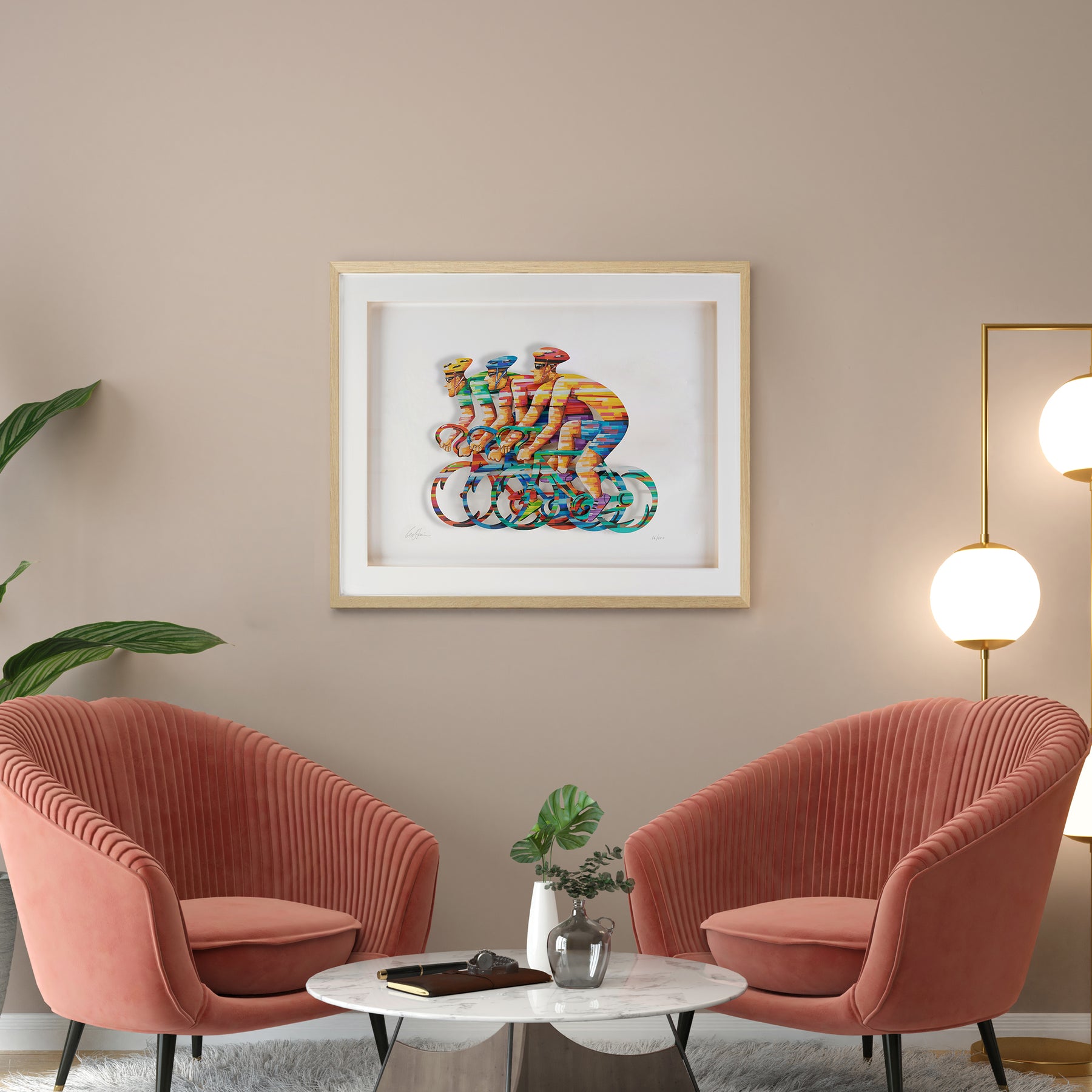 Papercut artwork depicting three people racing bicycles, each figure intricately cut and set against a clean white background, highlighting the delicate details and motion of the scene. The artwork is framed on the wall above two pink chairs.