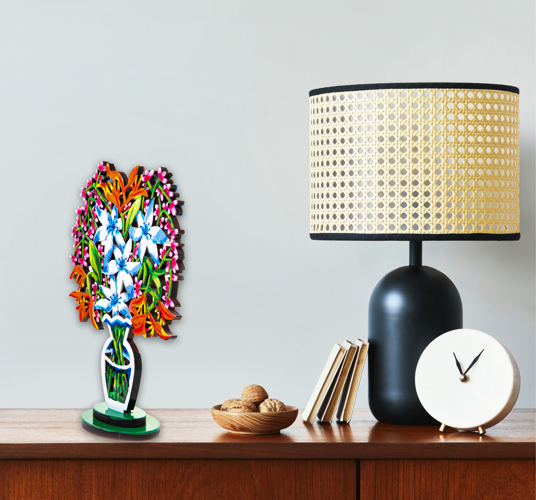 A a sculpture of a vibrant bouquet of blue and white flowers with orange and pink accents, displayed in a stylized, modern vase is seen placed on a table next to a black lamp.