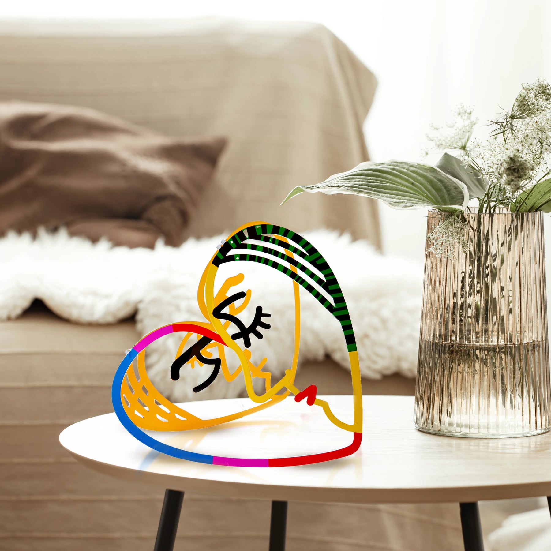 Small metal sculpture of a heart with two faces inside, placed on a coffee table next to a vase filled with fresh flowers. The heart sculpture adds a touch of artistry and warmth to the cozy setting.